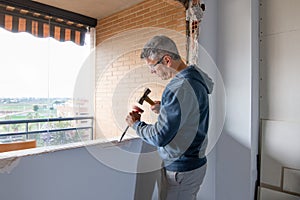 A bricklayer works on installing a new window in a home, chipping walls with a chisel and hammer and wearing protective glasses