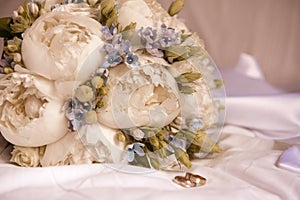 Bridal bouquet of peonies and blue flowers with  gold wedding rings on a white sheet