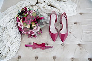 Bridal bouquet with red peonies, bow tie and red high-heeled shoes on a white pouffe, next to a knitted blanket