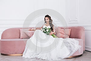 Bride in beautiful dress holding wedding bouquet and sitting on sofa indoors. Wedding day