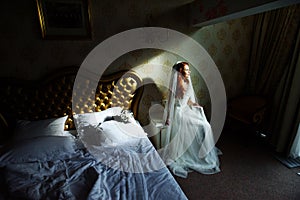 Beautiful redhair lady in elegant white wedding dress. Fashion portrait of model indoors. Beauty woman sitting near her bed