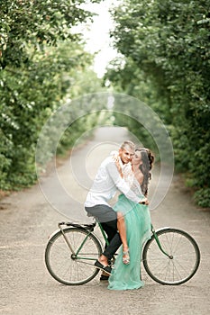 Bride and groom sit on bicycle on forest road, embrace and kiss.