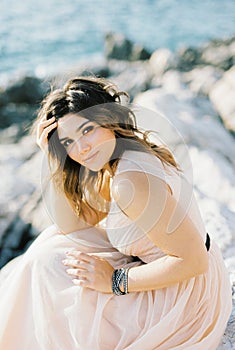 Bride in a beige dress sits on the rocks by the sea with her head propped up by her hand