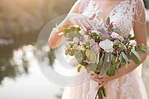 The bride in a beige wedding dress touching a lush bridal bouquet of lilac roses and a lot of greenery. Stylish wedding bouquet on