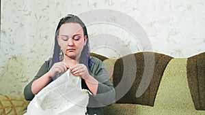 A bride with blue braids at home on a sofa sews her wedding dress by hand with a needle and thread