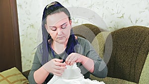 A bride with blue braids on a sofa sews her wedding dress by hand with a needle and thread