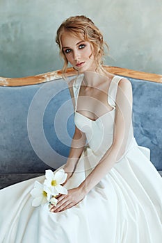 Bride in a chic long dress lying on the sofa couch. White wedding dress on the bride`s body. Beautiful light dress with a long he