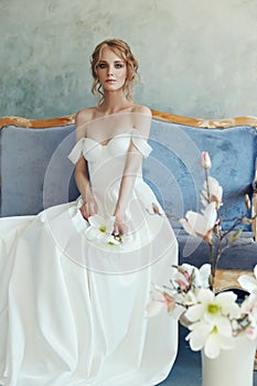 Bride in a chic long dress lying on the sofa couch. White wedding dress on the bride`s body. Beautiful light dress with a long he