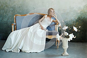 Bride in a chic long dress lying on the sofa couch. White wedding dress on the bride`s body. Beautiful light dress with a long he