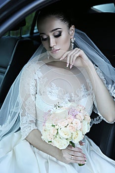 Bride with dark hair in luxurious wedding dress with bouquet of flowers sitting in car