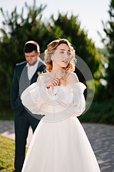 a bride in a delicate dress stands in front of the groom in the park.