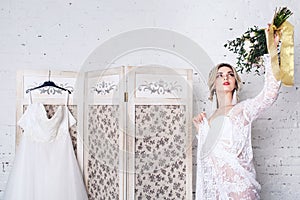 Bride, dressed in peignoir and underwear, stands near shield with wedding dress and holds bouquet in her hands.