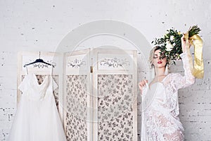 Bride, dressed in peignoir and underwear, stands near shield with wedding dress and holds bouquet in her hands.