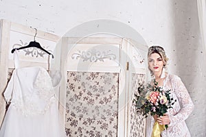 Bride, dressed in peignoir and underwear, stands near shield with wedding dress and holds bouquet in her hands.