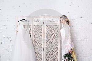 Bride, dressed in peignoir and underwear, stands near shield with wedding dress and holds bouquet in her hands.