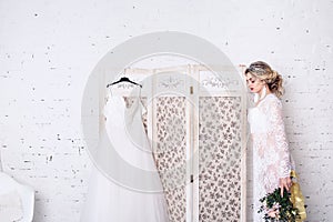 Bride, dressed in peignoir and underwear, stands near shield with wedding dress and holds bouquet in her hands.