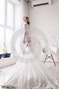 Bride, dressed in peignoir and underwear, stands near window and holds wedding dress in her hands. Morning of bride.