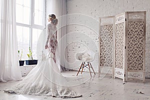 Bride, dressed in peignoir and underwear, stands near window and holds wedding dress in her hands. Morning of bride.