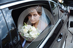 The bride is dressed in a wedding dress and veil, her head is draped with a lapel and sits in the car and admires the