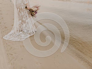 The bride is dressed in white lace wedding dress and holding wedding bouquet at the beach near the sea waves and glare.