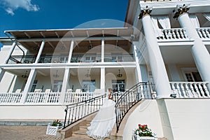 The bride goes up the stairs to the building with the columns. She holds a skirt of her white wedding dress, a bouquet