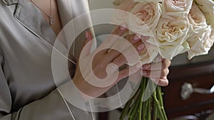 A bride in a gray robe holds a bouquet of white rose flowers in her hands