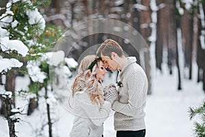 Bride and groom in beige knitted pullovers in snowy forest. Newlyweds is touching foreheads. Winter wedding