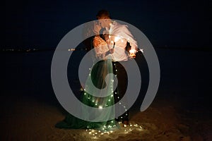 Bride and groom burn Bengal lights next to garland of glowing light bulbs at night.