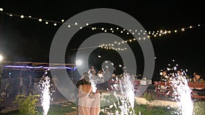 The bride and groom are dancing at a wedding party among fireworks, above them garlands with glowing bulbs