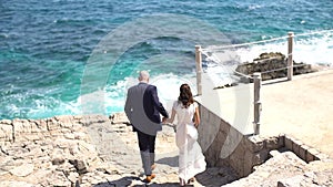 The bride and groom descend on the rocky seashore and hold hands