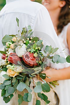 Bride and groom embracing, bride holding wedding bouquet of proteas, roses and eucalyptus in her hand