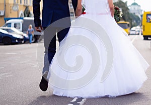 The bride and groom hold hands themselves while they walking on the road in city. Wedding in detail