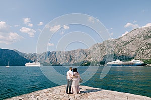 Bride and groom hug on the pier against the backdrop of liners and mountains. Back view