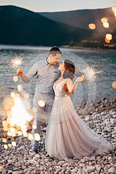 the bride and the groom hugging on the on the beach of the Mamula island and holding sparklers at the sunset