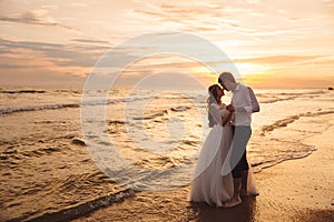 Bride and groom hugging and hold each other`s hands at beautiful sunset background. Newlyweds at wedding day on ocean