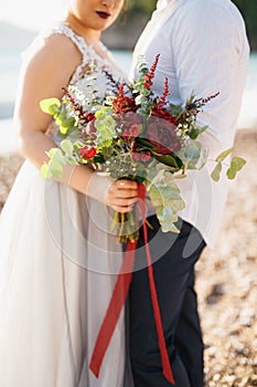 The bride and groom are hugging and holding wedding bouquet on the pebble beach by the sea