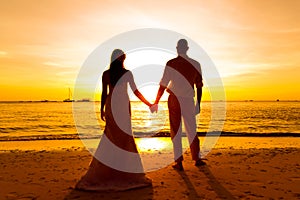 The bride and groom are kept hands on a tropical beach . Silhouette photo at sunset .