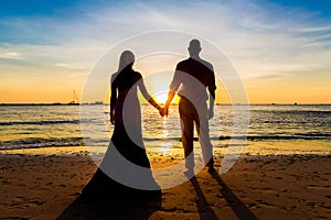 The bride and groom are kept hands on a tropical beach . Silhouette photo at sunset .