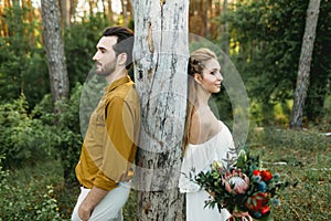 Bride and groom lean on the tree from different sides. Newlyweds are walking in the forest. Artwork