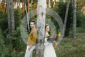 Bride and groom lean on the tree from different sides. Newlyweds are walking in the forest. Artwork