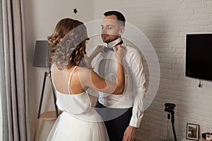 The bride and groom in the morning at the hotel on a wedding day. Dressing a newly-married couple Woman help fixing bowtie to groo