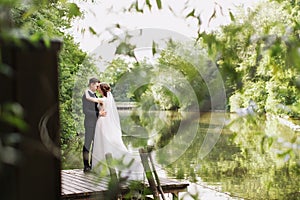 Bride and groom posing on the wooden pier near the pond among greenery. Young people embrace each other, and look at
