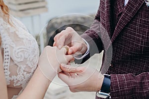 The bride and groom put on each other rings, a burgundy suit and a beige wedding dress, cloudy day, a ceremony by the sea