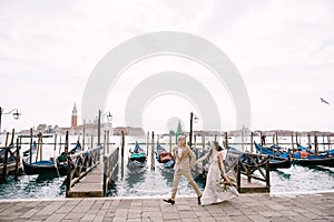 The bride and groom are run along the gondola pier, holding hand in Venice, near Piazza San Marco, overlooking San