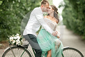 Bride and groom sit on bicycle on forest road, embrace and smile