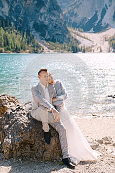 Bride and groom sit embracing against background of stones at the Lago di Braies in Italy. Destination wedding in Europe