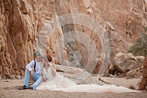 Bride and groom sit and smile in canyon on background of rocks.