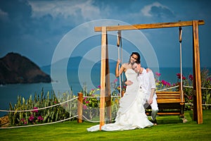 Bride and groom sit on a swing on the background of an incredibly beautiful landscape, ocean and mountains