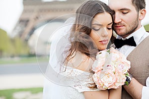 Bride and groom sitting on bench in park, holding hands of each other and bouquet. Groom holding his head on bride`s shoulder and