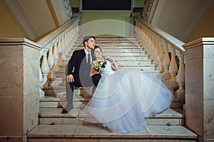 The bride and groom are sitting on a white marble staircase, holding hands and looking in the same direction